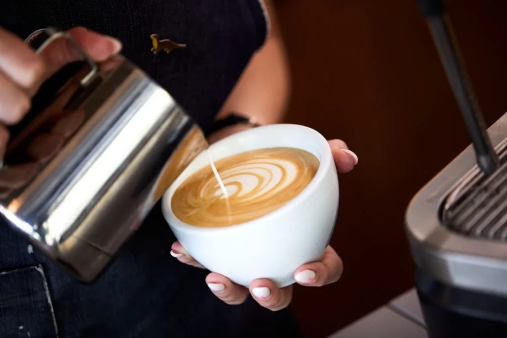 Barista tejet önt egy nagy csésze cappuccinóba, latte art mintát készítve.